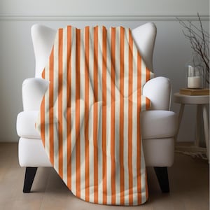 May include: A soft, orange and cream striped throw blanket draped over a white armchair. The blanket features vertical stripes and adds a pop of color to the neutral-toned room. The blanket is made of a plush material.