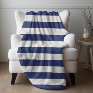 May include: A navy blue and white striped blanket draped over a white armchair. The blanket features thick, horizontal stripes. The chair is in a room with a neutral color palette, with a small side table and decorative elements visible.