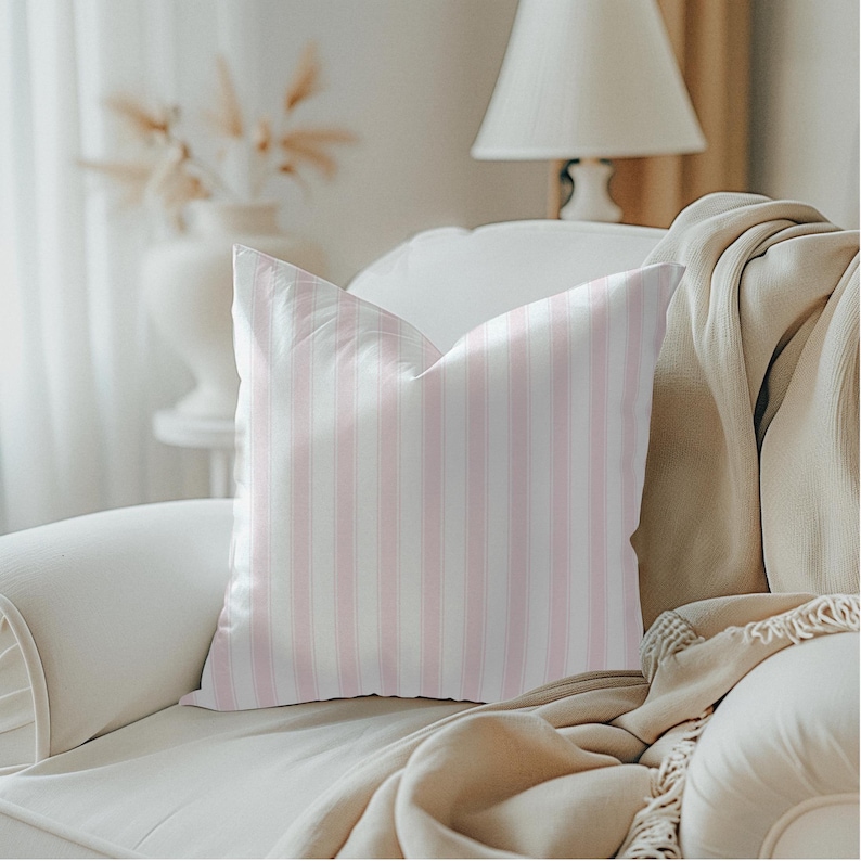 May include: A decorative throw pillow with vertical pink and white stripes. The pillow is placed on a white sofa, with a beige blanket draped over the arm. A lamp and vase are visible in the background.