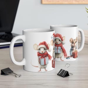 May include: White ceramic mugs featuring watercolor illustrations of mice in Santa hats and scarves, holding gifts. The mugs have a curved handle and are set on a light-colored surface, with office supplies in the foreground.