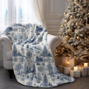 May include: A cozy, cream-colored throw blanket draped over a white armchair. The blanket features a blue and white winter scene with houses, trees, snowflakes, and a winding road. A decorated Christmas tree is in the background.