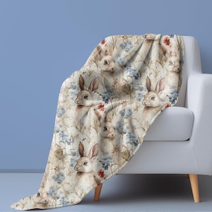 May include: A soft, plush throw blanket featuring a repeating pattern of watercolor bunnies and blue flowers on a cream background.