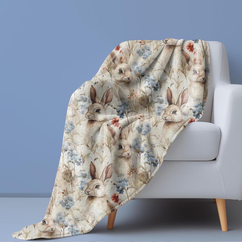 May include: A soft, plush throw blanket featuring a repeating pattern of watercolor bunnies and blue flowers on a cream background.