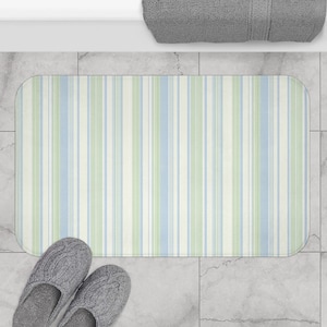 May include: A rectangular bath mat with vertical stripes in light blue, green, and off-white. The mat is placed on a tiled floor, with a pair of gray knit slippers at the bottom. A gray towel is visible at the top.