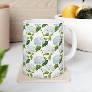 May include: White ceramic mug featuring a repeating pattern of blue and white hydrangea flowers with green leaves on a white background. The mug has a white handle and sits on a beige coaster. The mug is a kitchen and dining item.