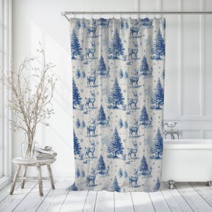 Op de afbeelding: Blauw-wit douchegordijn met een herhalend patroon van herten, dennenbomen en sneeuwvlokken. Het ontwerp heeft een toile-esthetiek, met de elementen gerangschikt in een winterse bosscène. Het gordijn hangt voor een wit bad.