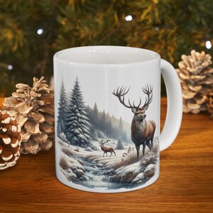 May include: White ceramic mug featuring a winter landscape with a majestic stag and a smaller deer near a stream. The design includes snow-covered trees and a forest backdrop, perfect for a cozy drink.