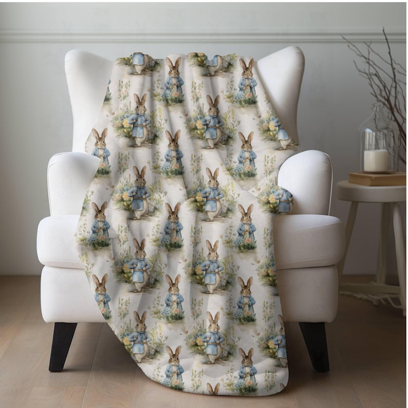 Bunny Rabbit Blanket. A white throw blanket with a repeating pattern of a brown rabbit wearing a blue jacket and holding a bunch of flowers. The rabbit is surrounded by green leaves and white flowers.