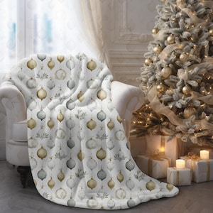 May include: A white throw blanket draped over a white armchair. The blanket features a repeating pattern of gold, gray, and white Christmas ornaments and sprigs. A decorated Christmas tree with gold ornaments and lights is in the background.