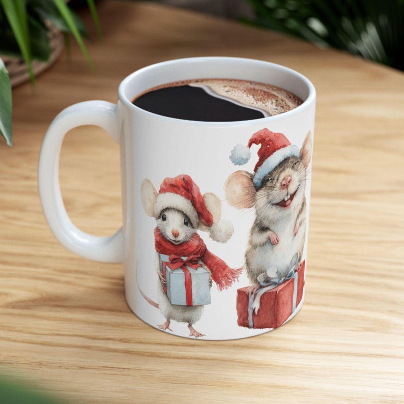 May include: White ceramic mug featuring two cartoon mice in Santa hats and scarves, holding gifts. One mouse is smiling. The mug is filled with dark liquid, likely coffee or tea. A festive holiday-themed design.