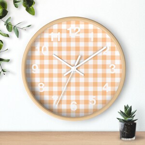 May include: A round wall clock with a light wood frame. The clock face features a white and orange gingham pattern with white numerals and hands. The clock is on a wooden surface.