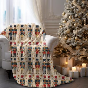 May include: A cream-colored throw blanket with a repeating pattern of vintage-style nutcracker soldiers in red, blue, and gold. The background features gold stars and small red and blue ornaments.