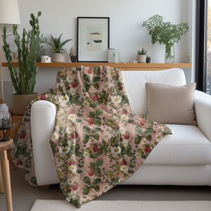 May include: A pink throw blanket with a floral pattern featuring strawberries, leaves, and white flowers. The blanket is draped over a white couch with a beige pillow.