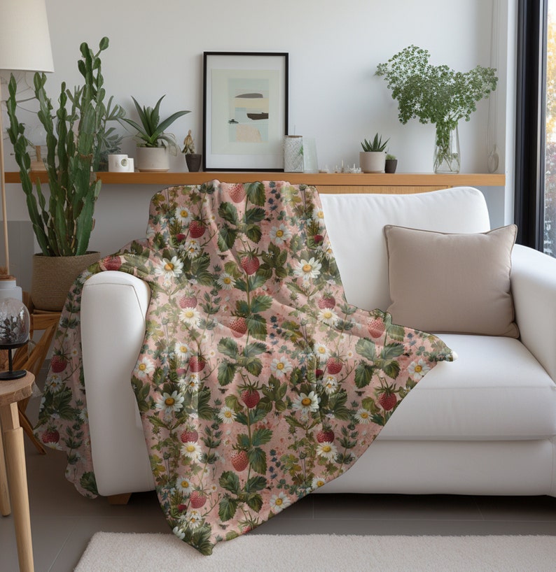 May include: A pink throw blanket with a floral pattern featuring strawberries, leaves, and white flowers. The blanket is draped over a white couch with a beige pillow.