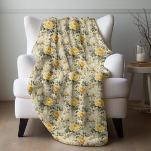 May include: A cream-colored throw blanket draped over a white armchair. The blanket features a repeating pattern of yellow roses and green leaves on a faded background. The blanket is made of a soft material and is suitable for a bed or sofa.