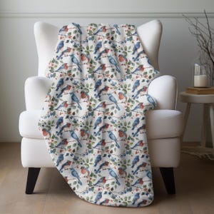 May include: A white throw blanket draped over a white armchair. The blanket features a repeating pattern of blue and red birds perched on branches with green leaves and berries. The blanket is made of a soft, comfortable material.