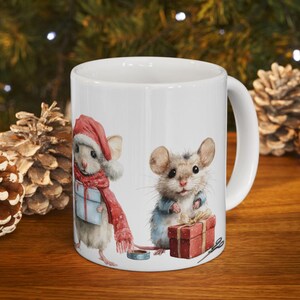 May include: White ceramic mug featuring two adorable watercolor mice in festive attire. One mouse wears a Santa hat and scarf, holding a gift. The other mouse is dressed in a blue jacket, with a red gift box. A charming holiday-themed mug.