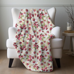 May include: A white throw blanket draped over a white armchair. The blanket features a repeating pattern of red raspberries with green leaves and yellow accents. The blanket adds a touch of nature to the room.