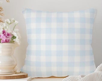May include: A square pillow with a light blue and white gingham pattern. The pillow is placed on a white surface next to a vase of flowers and a folded blanket. The pillow has a soft, inviting appearance.
