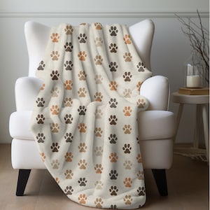 May include: A soft, plush throw blanket with a cream background and a repeating pattern of brown, orange, and beige paw prints.