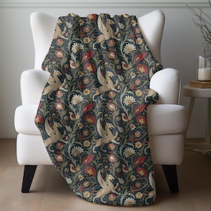 May include: A dark blue throw blanket draped over a white armchair. The blanket features a repeating pattern of gold winged creatures, red flowers, and cream-colored floral elements. The blanket is made of a soft material.