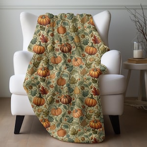 May include: A cozy throw blanket draped over a white armchair. The blanket features a repeating pattern of orange pumpkins, green leaves, and red foliage on a cream background. The design evokes a fall or harvest theme.