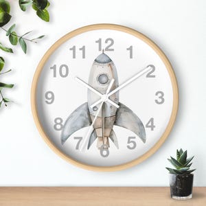 May include: A round wall clock with a light wood frame. The clock face is white with gray numbers and a watercolor illustration of a rocket ship. The clock hands are white.