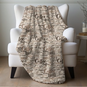 May include: A soft, beige throw blanket with a repeating pattern of the word "Coffee" in various shades of brown. The blanket is draped over a white armchair.
