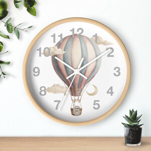 May include: A round wall clock with a light wood frame. The clock face features a watercolor illustration of a hot air balloon in shades of pink, blue, and white, with clouds and a crescent moon. The numbers are gray.