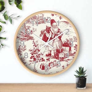 May include: A round wall clock with a light wood frame. The clock face features a red and white toile design with a Santa Claus illustration, surrounded by gifts and a winter scene. Silver numerals mark the hours.