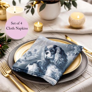 May include: Set of four blue and white cloth napkins featuring a Cavalier King Charles Spaniel dog illustration. The napkins are folded and placed on a gold and gray dinner plate.