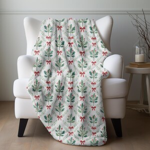 May include: A white throw blanket draped over a white armchair. The blanket features a repeating pattern of green holly branches, red bows, and red berries. The blanket is made of a soft, plush material.