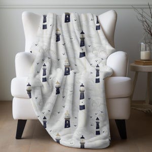 May include: A white throw blanket draped over a white armchair. The blanket features a repeating pattern of navy blue and gold lighthouses, stylized waves, and small black birds. The blanket has a soft, plush texture.