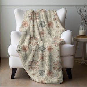 May include: A beige throw blanket draped over a white armchair. The blanket features a repeating pattern of stylized starbursts in shades of red, gray, and brown. The blanket is made of a soft material and is suitable for home decor.