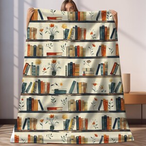 May include: A cream-colored throw blanket with a repeating pattern of bookshelves filled with colorful books and small floral arrangements. The bookshelves are brown with white shelves.