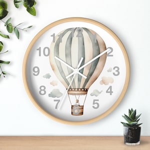May include: A white clock with a wooden frame features a watercolor illustration of a hot air balloon with blue and beige stripes. The clock face has silver numbers and hands.