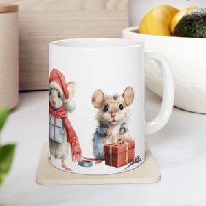 May include: White ceramic mug featuring watercolor illustrations of two mice. One mouse wears a Santa hat and red scarf, holding a blue gift. The other mouse wears a blue jacket and holds a red gift box. A festive holiday theme.