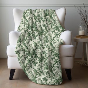 May include: A cream-colored throw blanket draped over a white armchair. The blanket features a repeating pattern of green owls perched in a leafy tree design. The owls and foliage are rendered in a detailed, vintage style, creating a cozy, nature-inspired aesthetic.