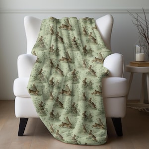 May include: A soft, light green throw blanket draped over a white armchair. The blanket features a repeating pattern of brown rabbits in various poses amidst foliage. The design has a vintage, illustrated style, perfect for adding a touch of nature to any room.