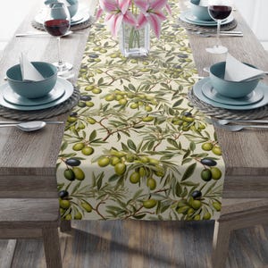 May include: A beige table runner with a repeating pattern of olive branches and green olives. The runner is on a wooden table with two place settings on each side.