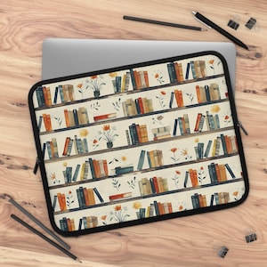 May include: Laptop sleeve featuring a watercolor design of bookshelves with books in various colors, potted plants, and flowers. The sleeve has a black zipper and trim, and is resting on a wooden surface with a laptop and pens.