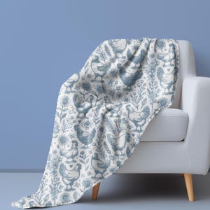 May include: A white throw blanket draped over a white armchair. The blanket features a repeating pattern of blue chickens, flowers, and foliage. The blanket is made of a soft material and is suitable for a living room or bedroom.