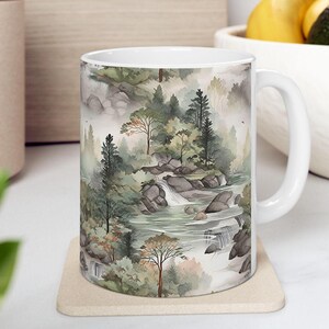 May include: A white ceramic mug featuring a wrap-around watercolor illustration of a misty forest landscape. The design depicts evergreen trees, rocky terrain, and cascading waterfalls flowing into a stream, rendered in muted shades of green and gray.