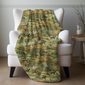May include: A throw blanket draped over a white armchair. The blanket features a repeating pattern of riders on horseback, foxes, and rolling green hills. The color palette includes shades of green, brown, and yellow.