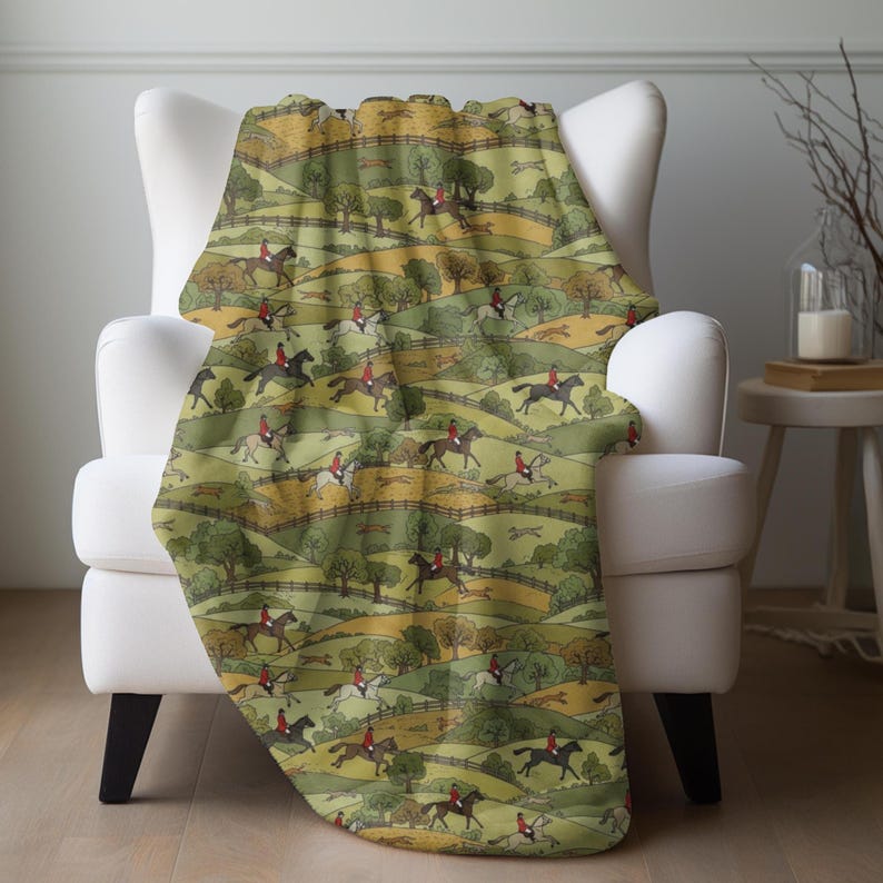 May include: A throw blanket draped over a white armchair. The blanket features a repeating pattern of riders on horseback, foxes, and rolling green hills. The color palette includes shades of green, brown, and yellow.