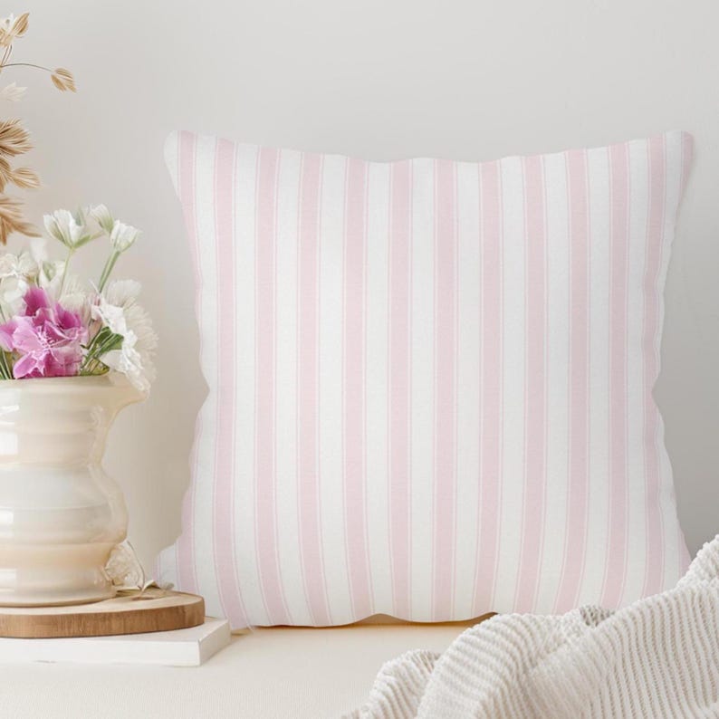 May include: A square decorative pillow with a vertical stripe pattern in soft pink and white. The pillow is placed on a white surface, with a vase of flowers and a folded blanket in the background.