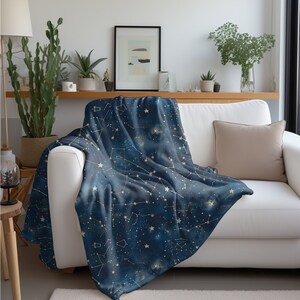 以下が含まれることがあります： 金色の星と線で構成された星座模様が施された、柔らかなブルーの毛布。白いソファにベージュのクッションを添えて、毛布がかけられています。