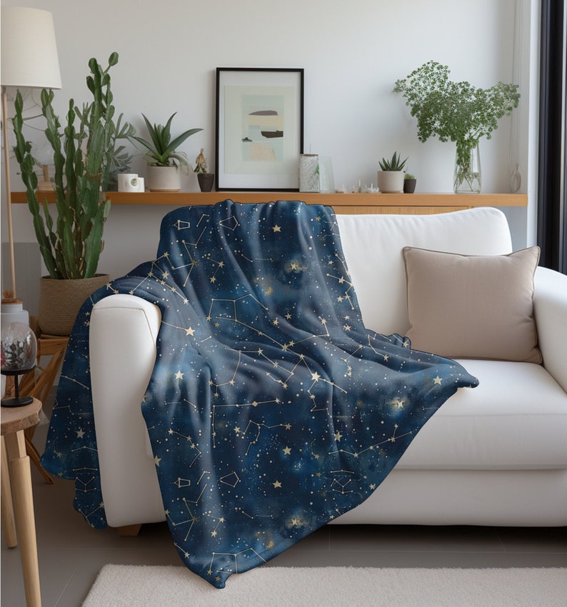 以下が含まれることがあります： 金色の星と線で構成された星座模様が施された、柔らかなブルーの毛布。白いソファにベージュのクッションを添えて、毛布がかけられています。