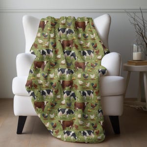 May include: A green throw blanket draped over a white armchair. The blanket features a repeating pattern of brown and black cows, chickens, and roosters on a green background. The blanket is made of a soft material.