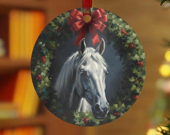 May include: Round Christmas ornament featuring a white horse's head within a wreath of green leaves and red berries. A red ribbon and bow are at the top. The ornament is hanging from a red ribbon.